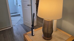Un couloir avec un plancher en bois, une porte blanche, une lampe avec un abat-jour beige et un vase contenant des tiges noires.