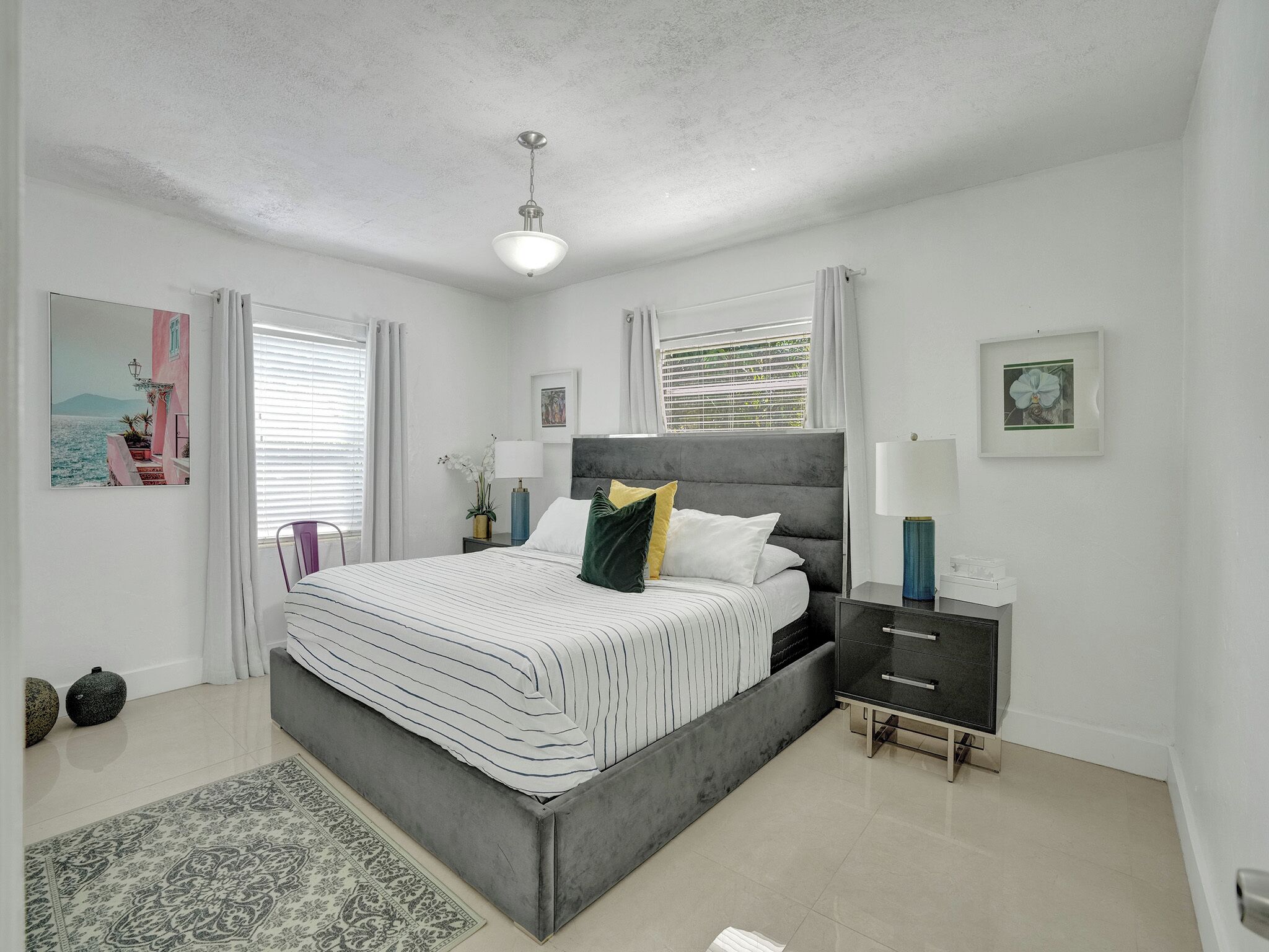 A bedroom with a bed, bedside tables, a nightstand, a lamp, a window with curtains, and a painting on the wall.