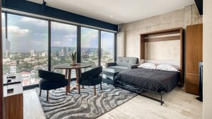 A modern bedroom with a large bed, a sofa, a dining table, and a view of the city.