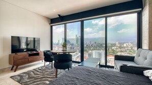 A modern bedroom with a large window, a flat-screen TV, a sofa, a dining table, and a city view.
