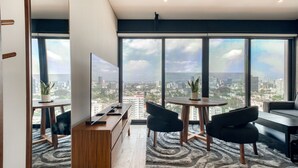 A modern living room with a large window, a flat-screen TV, a wooden coffee table, and two black chairs.