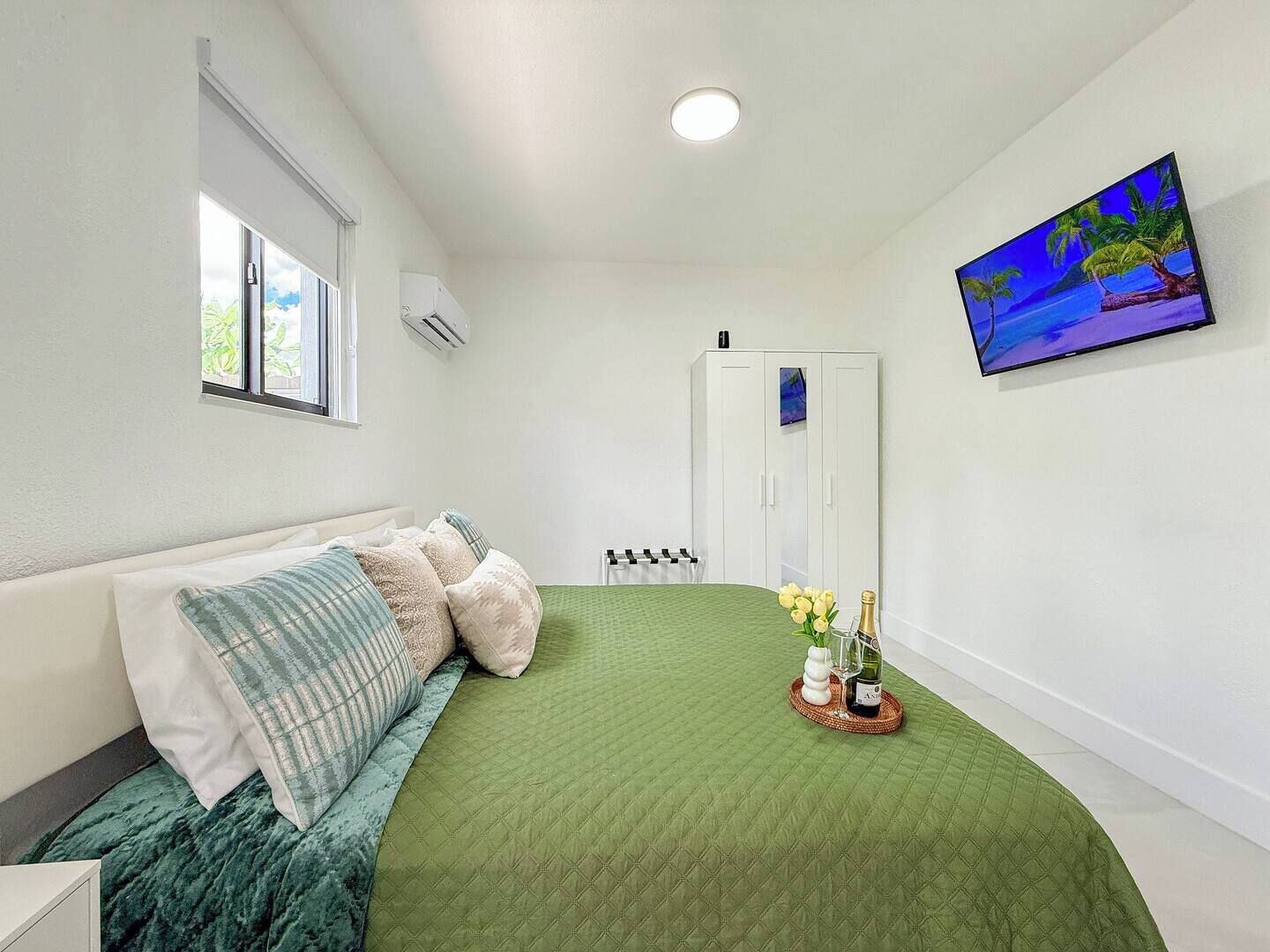 A bedroom with a green quilted bedspread, a white wardrobe, a painting of a beach scene, and a window with a view of greenery.