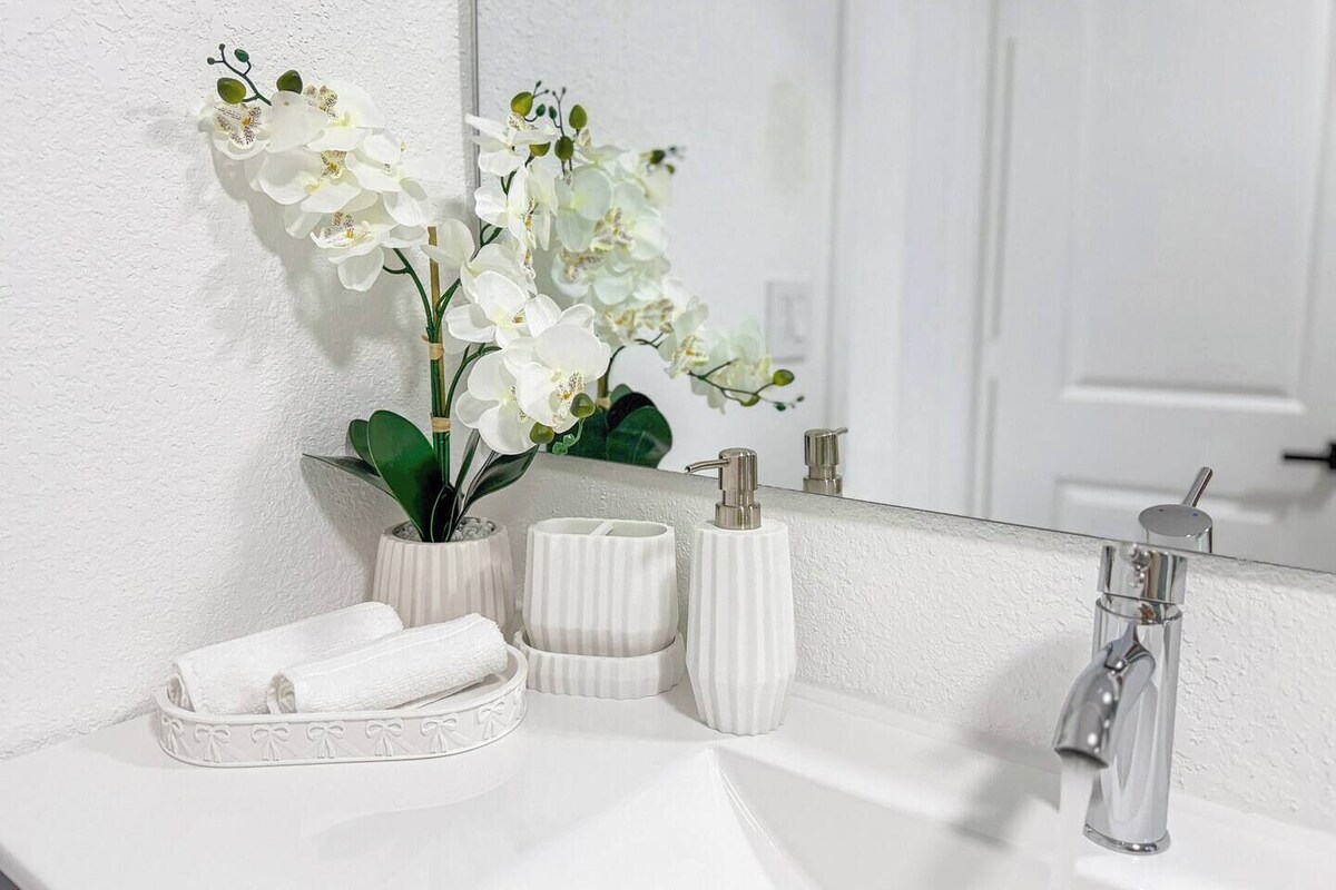 A bathroom with a white sink, a mirror, and a toilet. There are white towels and a white vase with flowers on the sink.