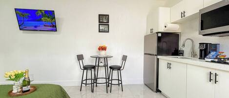 A compact kitchen with white cabinets, a stainless steel refrigerator, a microwave, and a coffee maker. There is a small dining area with a round table and four chairs. A flat-screen TV is mounted on the wall above the dining area.