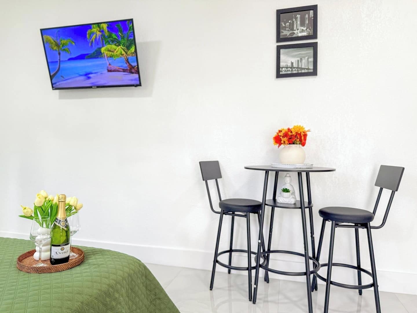 Habitación de hotel con una cama, una mesita pequeña con dos sillas, un televisor montado en la pared y dos cuadros enmarcados.
