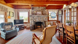 A cozy living room with a stone fireplace, wooden ceiling, and a dining area with a table and chairs. - Quiet Oceanfront Cottage with Dock (Boothbay Harbor)