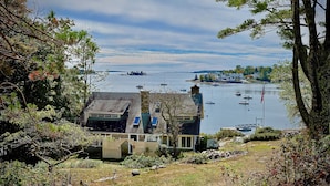 A house with a sloped roof, a fenced yard, and a view of a calm lake with boats. - Quiet Oceanfront Cottage with Dock (Boothbay Harbor)
