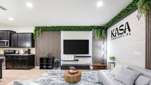Ein modernes Wohnzimmer mit Flachbildfernseher, einer Couch, einem Couchtisch und einer Küchenzeile mit dunklen Schränken und einer Mikrowelle.