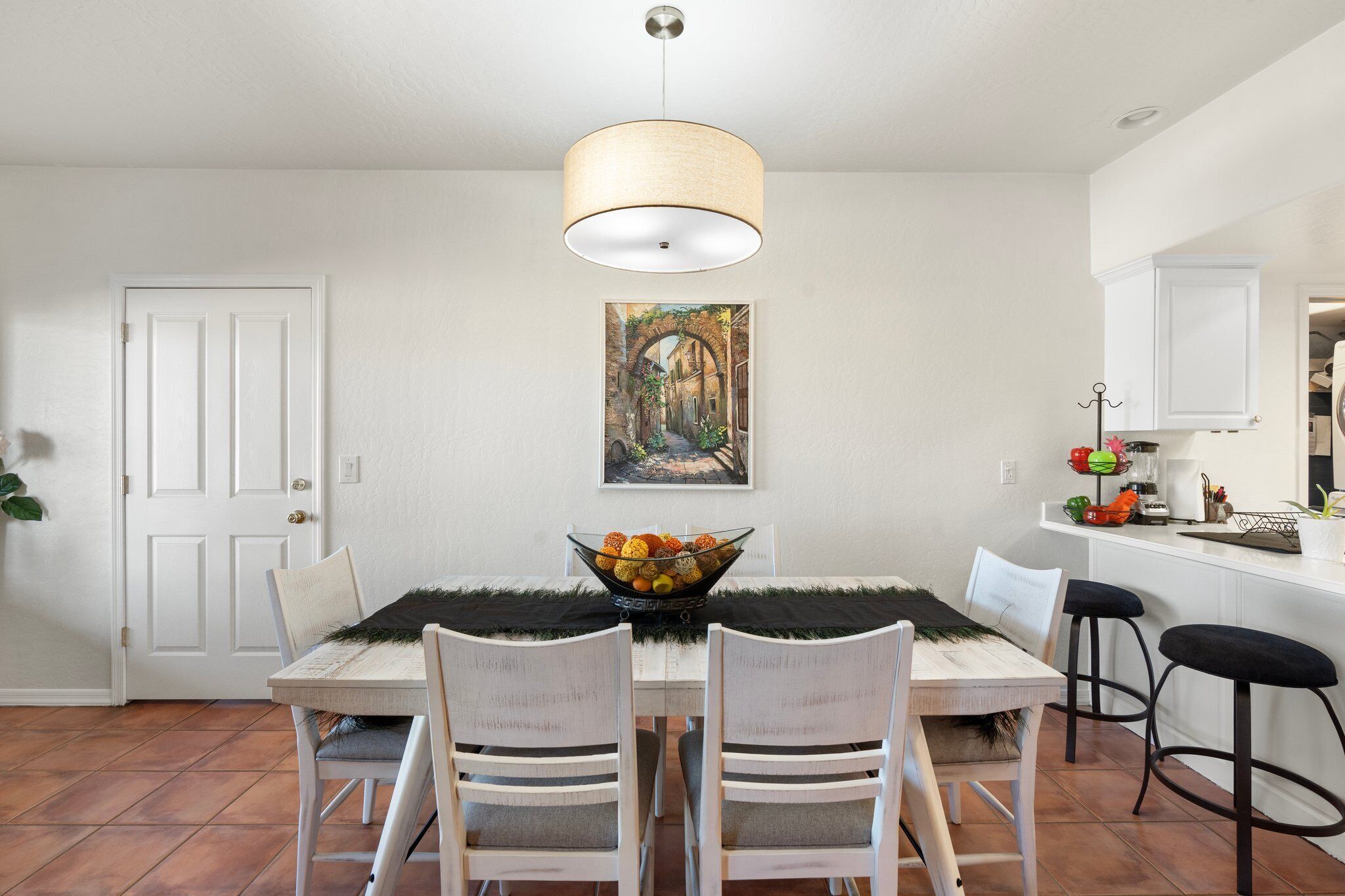 A dining area with a white table, two chairs, a bowl of fruit, and a hanging light fixture.