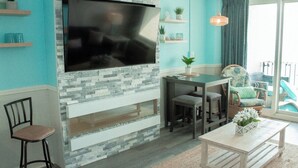 A living room with a stone fireplace, a flat-screen TV mounted above it, a dining area with a table and chairs, and a balcony with a view. - Myrtle Beach Quiet Coastal Getaway | Great Ocean & Coast Views (Myrtle Beach)