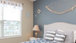 A bedroom with a bed, a nightstand, a lamp, and wall decorations including a life preserver and a net. - Heatherston 1063 Walk to Tee Time Daily overlooking the pool area! (Myrtle Beach)