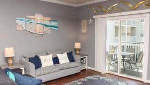 A living room with a grey sofa, a blue and white pillow, a wooden floor, a patterned rug, and a balcony with outdoor furniture. - Heatherston 1063 Walk to Tee Time Daily overlooking the pool area! (Myrtle Beach)