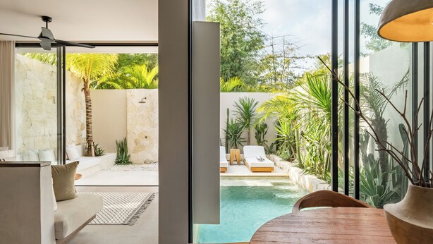 Un intérieur moderne avec un espace piscine visible à travers de grandes fenêtres, comprenant un canapé, une table à manger et un ventilateur de plafond.