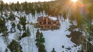 A large, two-story log cabin with a wrap-around porch, surrounded by a forested area with snow on the ground. - Views | Hot Tub | Pet Friendly | Sunrise Ridge (Angel Fire)