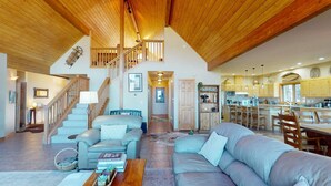 A spacious living area with a high ceiling, a staircase leading to an upper level, a kitchen with wooden cabinets, and a dining area with chairs. - Views | Hot Tub | Pet Friendly | Sunrise Ridge (Angel Fire)