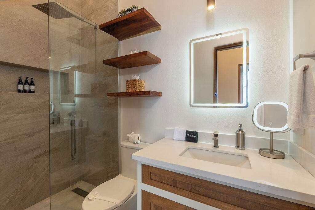 Ein modernes Badezimmer mit einer Duschkabine aus Glas, einem weißen Waschbecken mit Spiegel und Holzregalen über der Toilette.