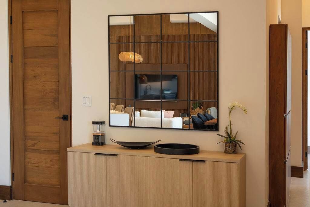 Un salon moderne avec un grand miroir qui reflète l’intérieur, un meuble en bois orné d’un vase et d’une plante, et une porte dotée d’un panneau texturé.