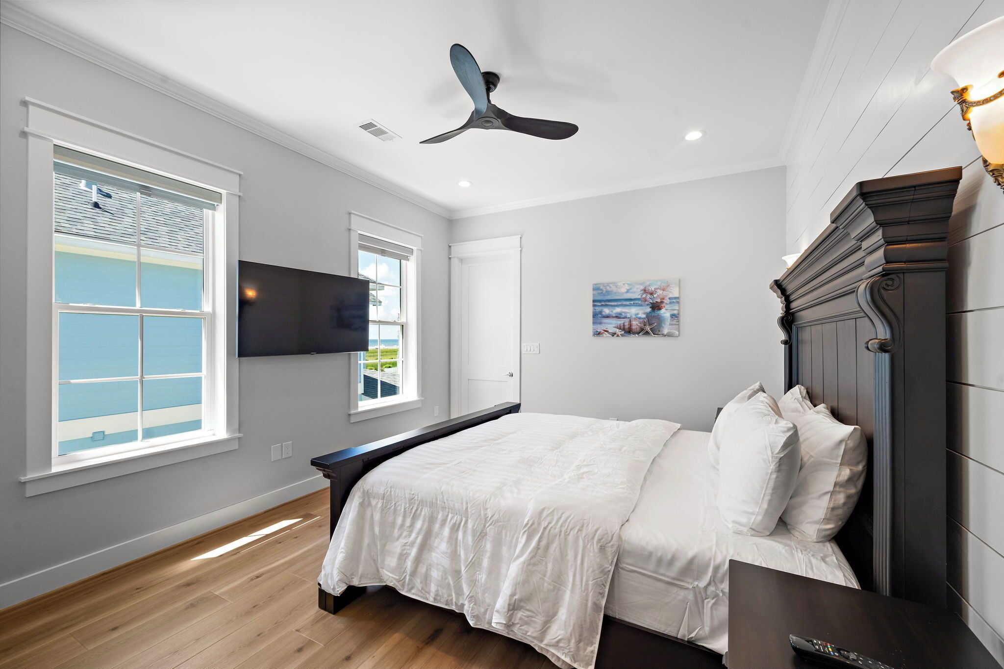 Une chambre à coucher avec un lit, un ventilateur de plafond, un téléviseur fixé au mur et une fenêtre avec des stores.