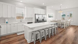 A modern kitchen with white cabinetry, a large island with bar stools, stainless steel appliances, and a dining area with a white table and chairs. - Ellis Retreat with LSV, Arcade, and Billiards (Miramar Beach)