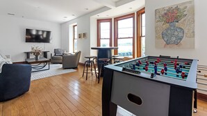 Un salon moderne avec un baby-foot, des sièges confortables, un téléviseur et de grandes fenêtres.