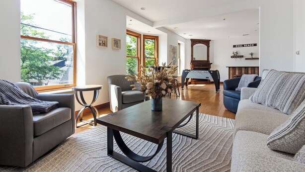 Un salon spacieux avec un canapé gris, une table basse et un fauteuil bleu. Une grande fenêtre laisse entrer la lumière naturelle et offre une vue sur les arbres.