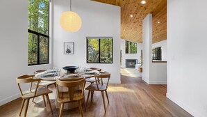 A spacious dining area with a round wooden table and chairs, large windows offering views of trees, and a wooden ceiling.