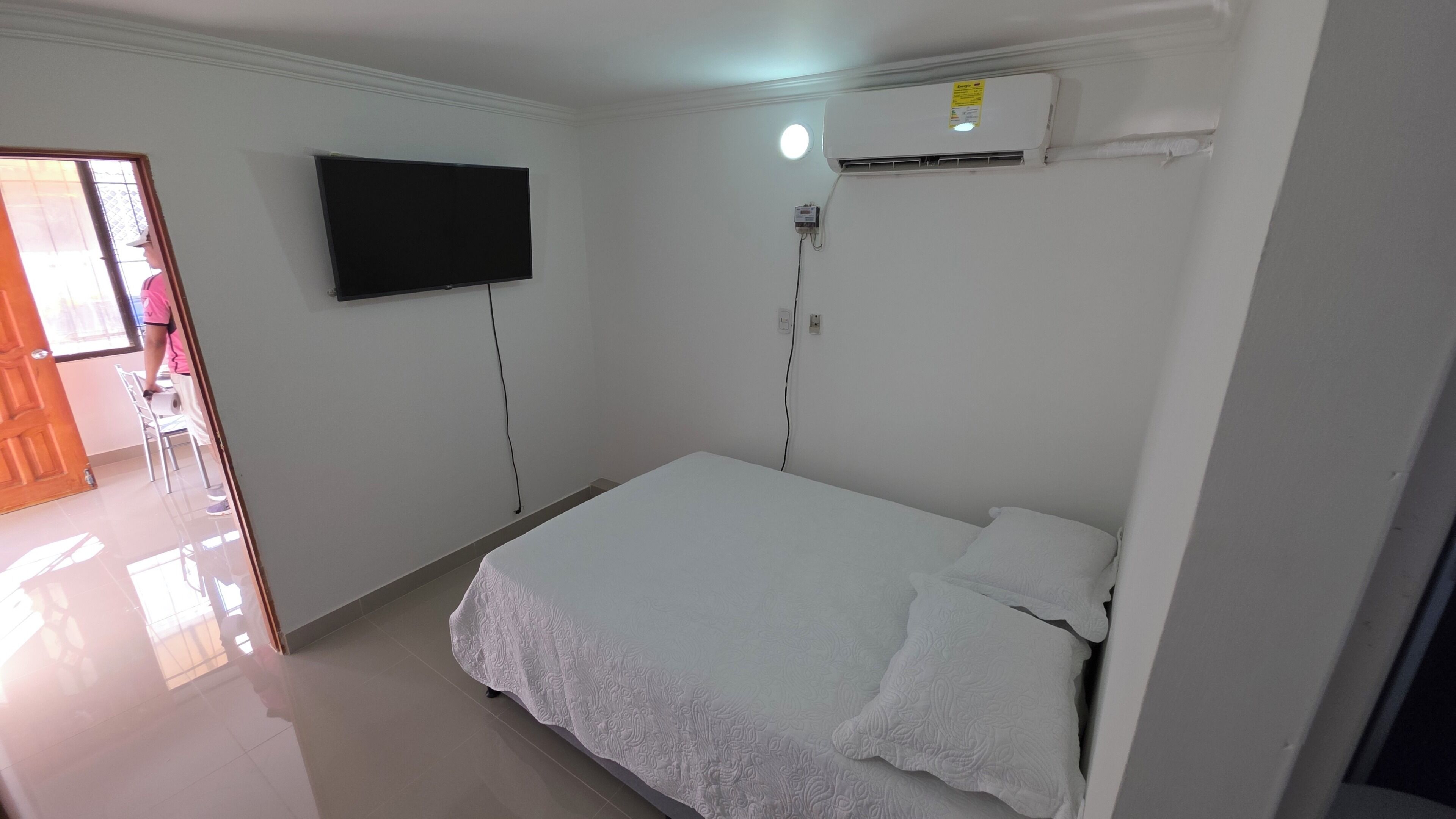 Une chambre à coucher comprenant un lit, un téléviseur fixé au mur, un appareil de climatisation et une porte donnant sur une autre pièce.