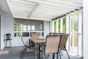A covered patio with a table and chairs, large windows, and a view of greenery.