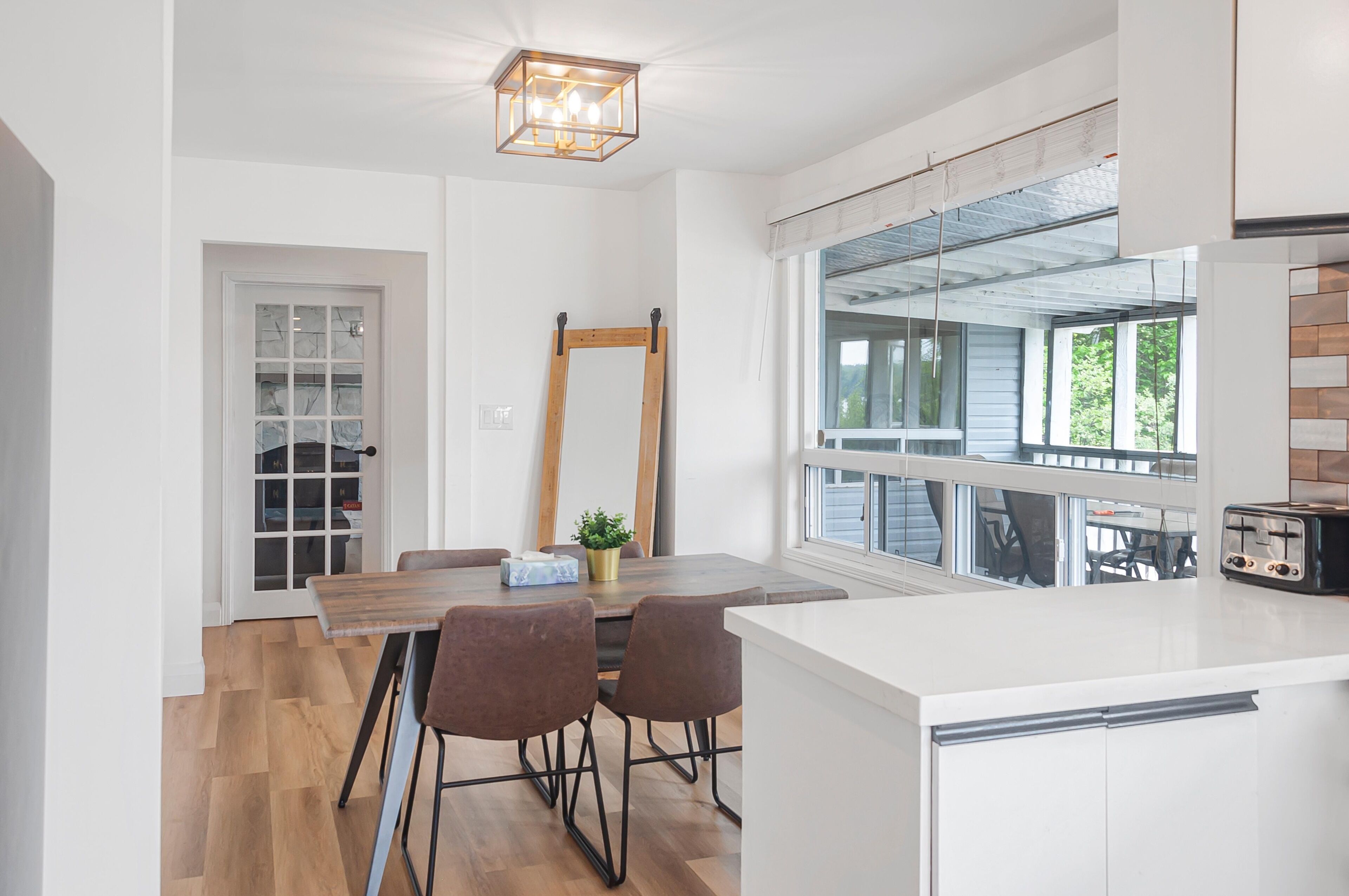 A modern kitchen with a wooden dining table, two chairs, a hanging light fixture, and a large window with a view of the outdoors.