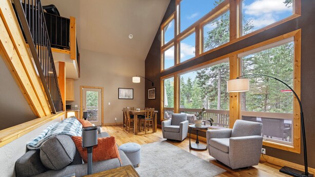A spacious living area with a high ceiling, large windows, a staircase, and a cozy seating arrangement. - New! Hot tub, Fire Pit, Foosball Table! (Bigfork)