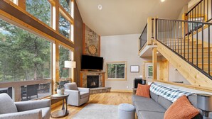 A spacious living room with a fireplace, large windows, and a wooden staircase. - New! Hot tub, Fire Pit, Foosball Table! (Bigfork)