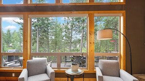 A room with two grey armchairs, a small round table with a potted plant, and a floor lamp. Large windows offer a view of a deck and surrounding trees. - New! Hot tub, Fire Pit, Foosball Table! (Bigfork)