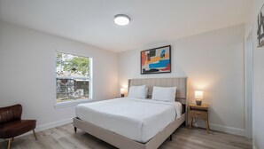 Ein Schlafzimmer mit einem Bett, einem Nachttisch, einer Lampe, einem Sessel und einem Fenster mit Jalousien.