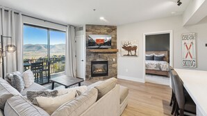 A modern living room with a fireplace, a large window with a mountain view, a dining area, and a bedroom with a ski-themed wall art. - Luxury 2 Bedroom Condo at The Mason Pool, Free Ski Shuttle, AC (Heber City)