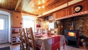 A rustic kitchen with a wood-burning stove, wooden chairs, a dining table with a red tablecloth, and a stone wall. - Chez l'Toine et la Pauline (SAINT ROMAIN)