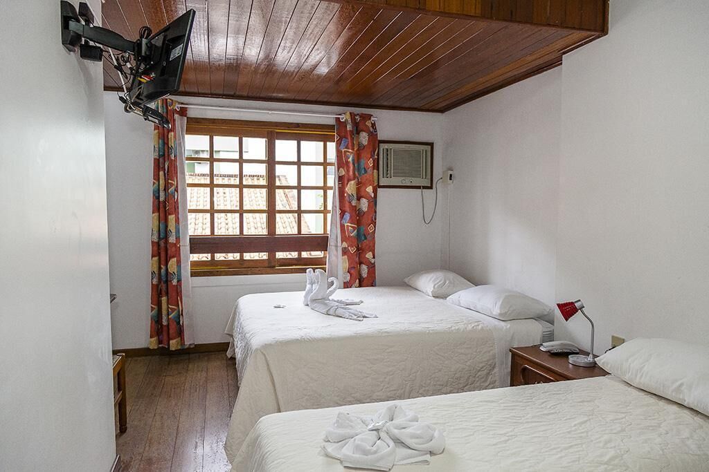 Quarto de hotel com duas camas, uma TV presa na parede, uma janela com cortinas e uma mesinha de cabeceira com abajur.