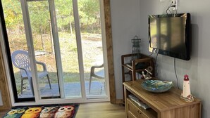 A room with a wooden desk, a television mounted on the wall, a small trash bin, and a view through a sliding glass door to an outdoor area with chairs.