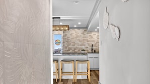 A modern kitchen with a central island, wooden bar stools, and decorative wall art. - Oceanfront 3BR Dolphin Run condo with balcony and pool. (Virginia Beach)