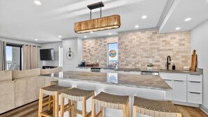 A modern kitchen with a large island, bar stools, a sofa, and a TV. - Oceanfront 3BR Dolphin Run condo with balcony and pool. (Virginia Beach)