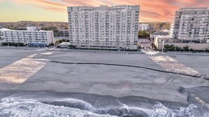 A beach with waves, a large building with many windows, and a sunset in the background. - Oceanfront 3BR Dolphin Run condo with balcony and pool. (Virginia Beach)