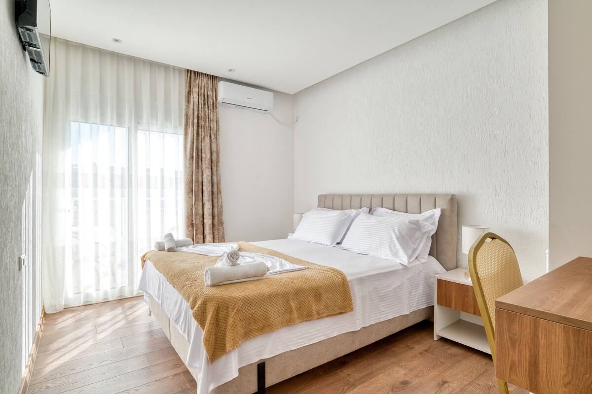 A modern hotel room with a bed, a wooden nightstand, a chair, and a window with curtains.