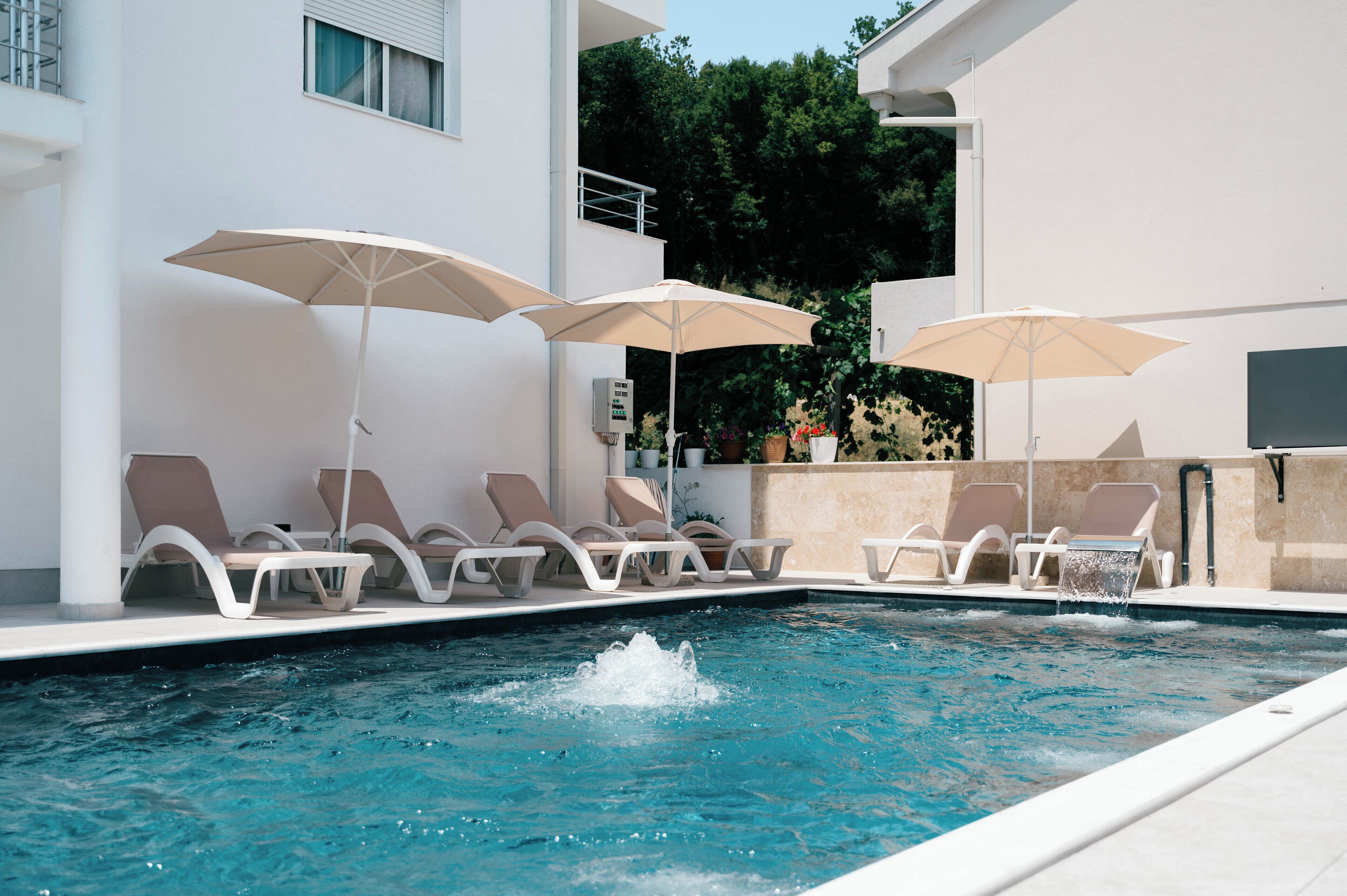 A pool area with lounge chairs and umbrellas, a water feature, and a building with a balcony.