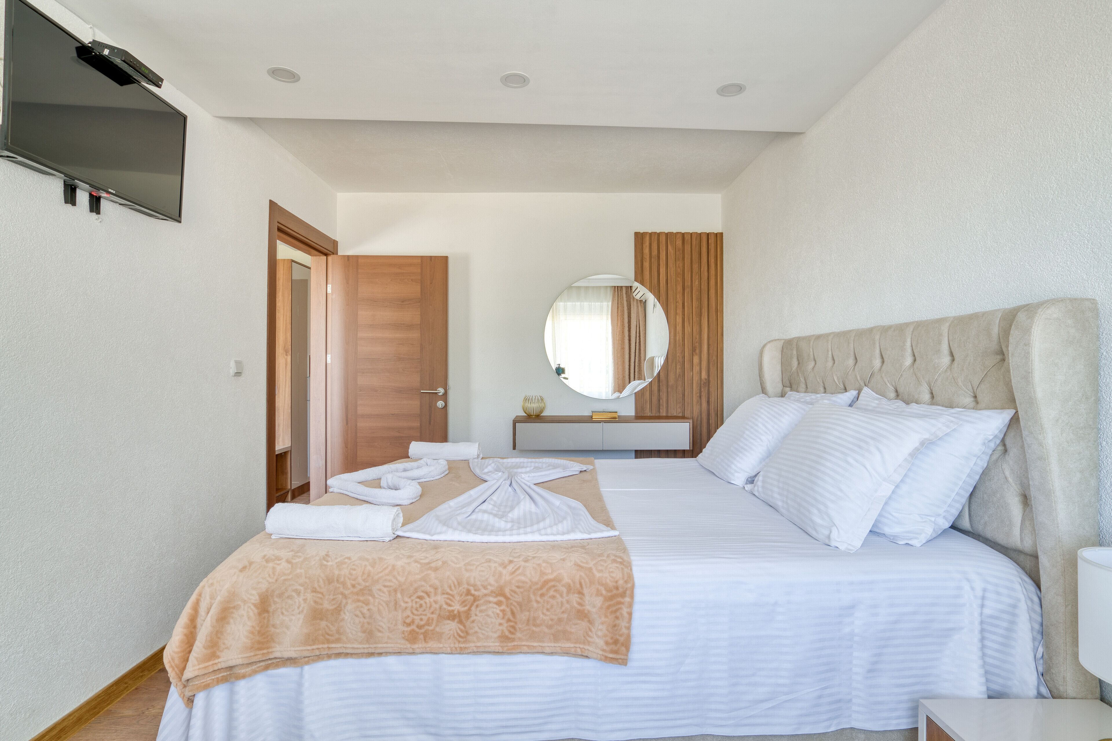 A hotel room with a bed, a television mounted on the wall, a round mirror, and a bedside table.