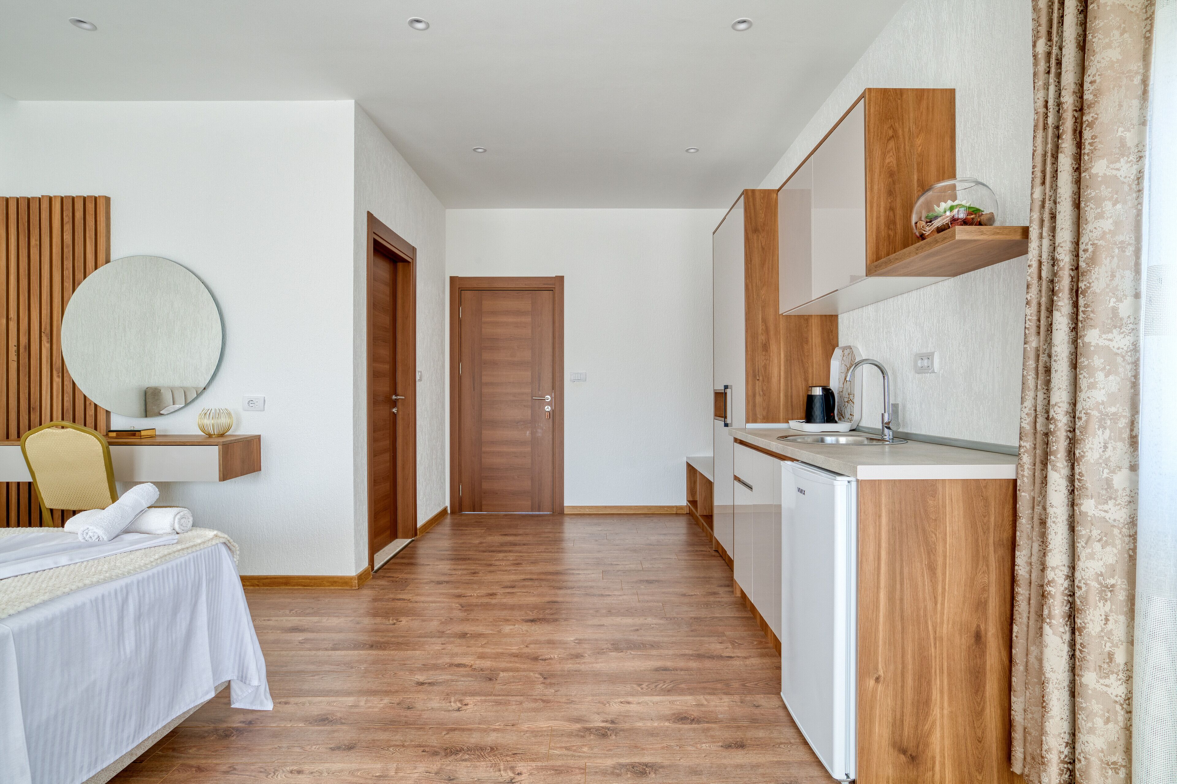 Una habitación de hotel moderna con suelo de madera, una cama con sábanas blancas, un espejo redondo, una mesita con un jarrón, una estantería de madera con una cafetera y una puerta que da a otra habitación.