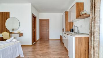 Una habitación de hotel moderna con suelo de madera, una cama con sábanas blancas, un espejo redondo, una mesita con un jarrón, una estantería de madera con una cafetera y una puerta que da a otra habitación.