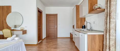 Una habitación de hotel moderna con suelo de madera, una cama con sábanas blancas, un espejo redondo, una mesita con un jarrón, una estantería de madera con una cafetera y una puerta que da a otra habitación.