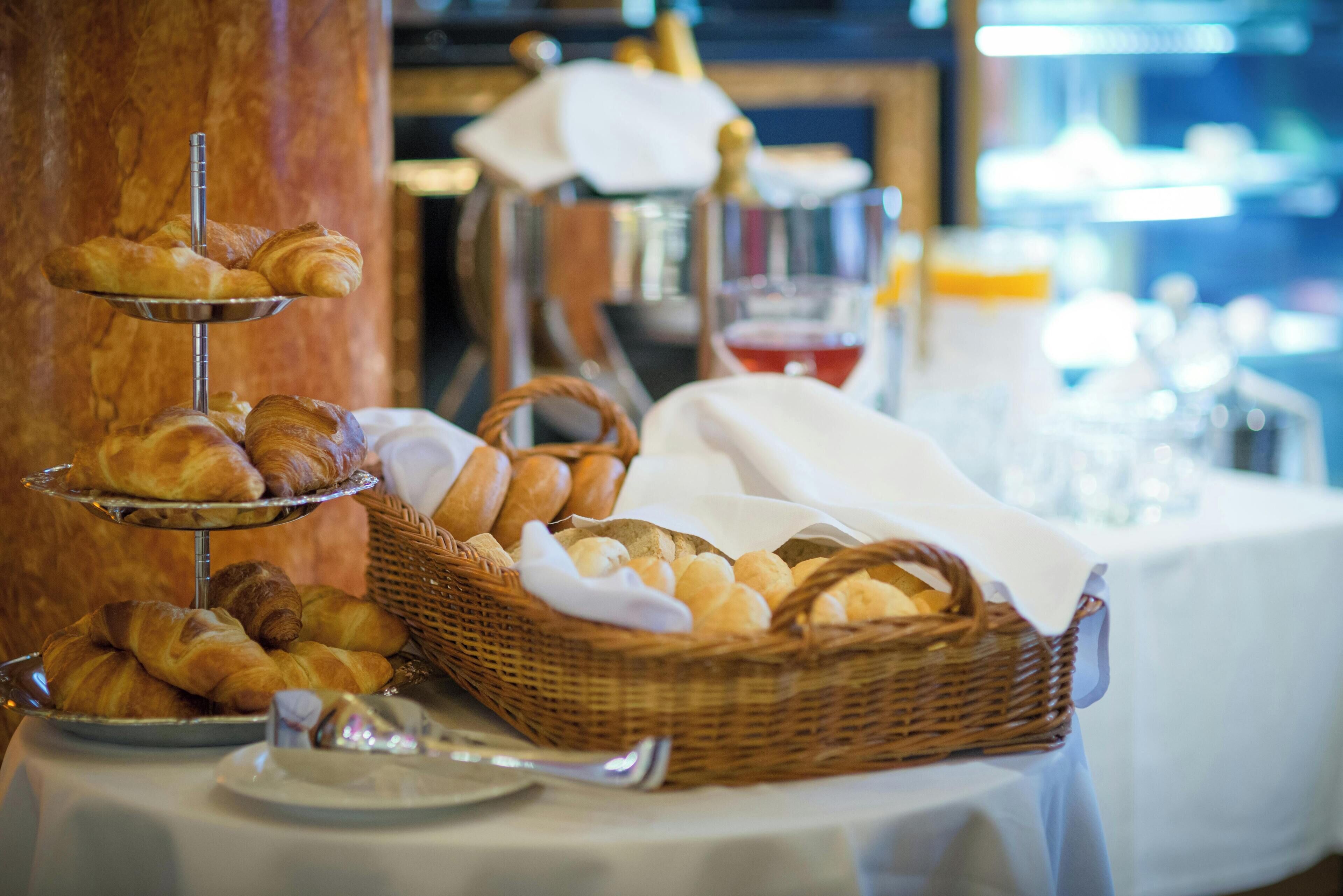 Una mesa de buffet con cruasanes y pan, una cesta de pan y una botella de champán al fondo.