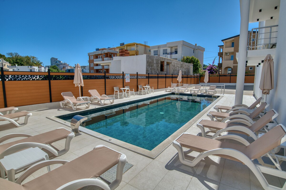 A poolside area with lounge chairs and umbrellas, surrounded by a wooden fence and buildings in the background.