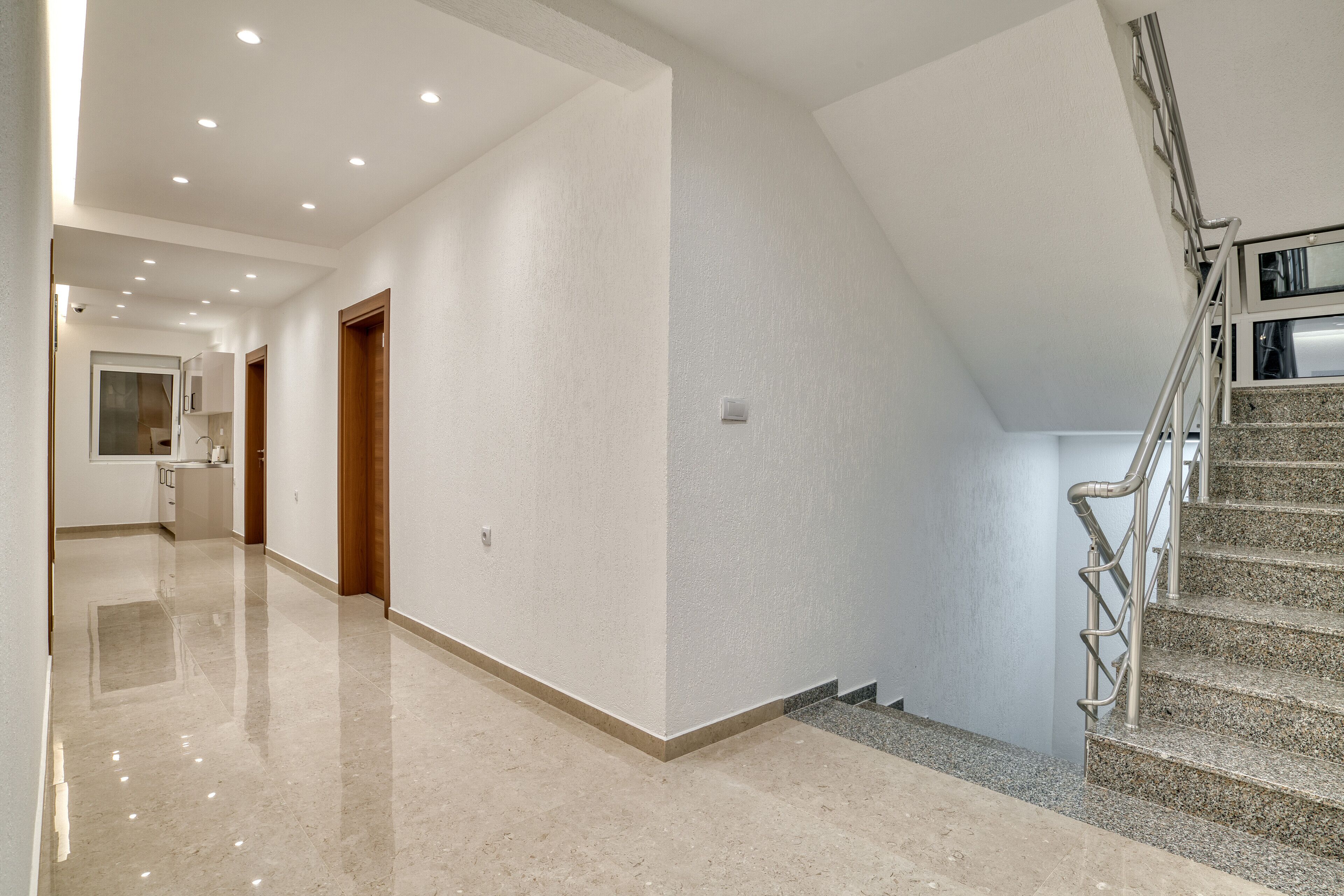 Un hall d’entrée spacieux avec un plancher poli, un escalier doté de rampes en métal et un coin cuisine visible au fond.