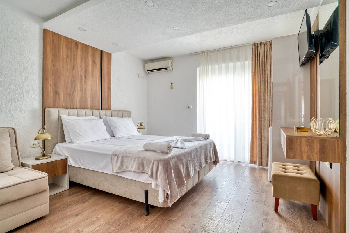 A modern hotel room with a large bed, a wooden accent wall, a flat-screen TV mounted on the wall, and a small seating area.